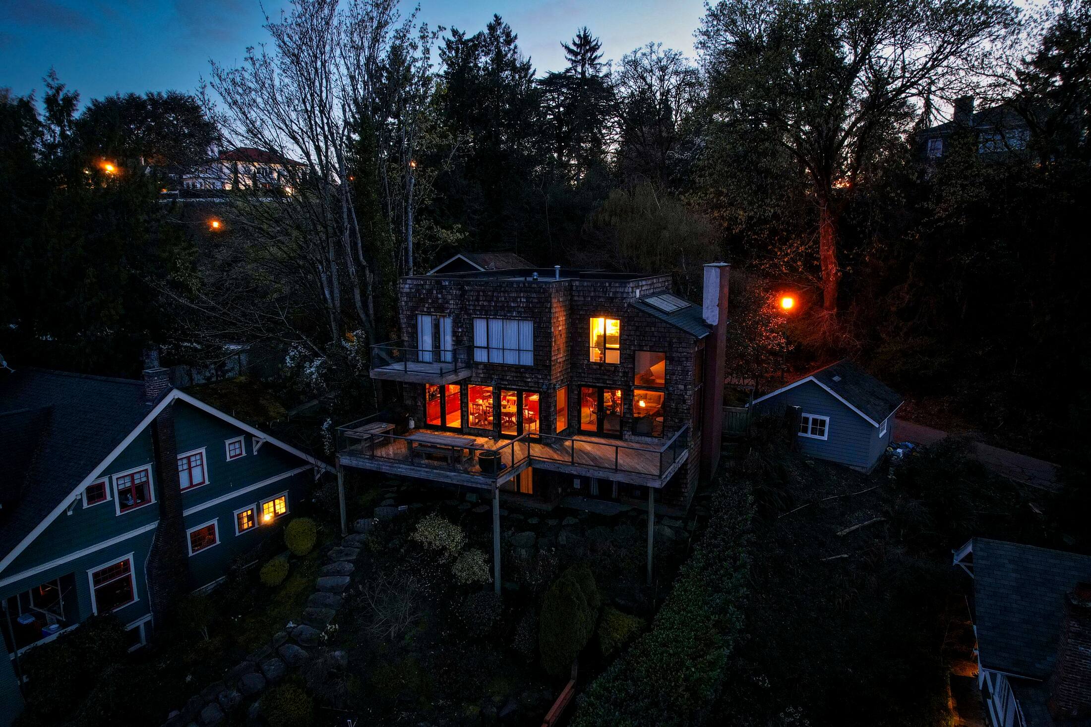 Home glowing warmly at dusk beneath tall evergreen trees